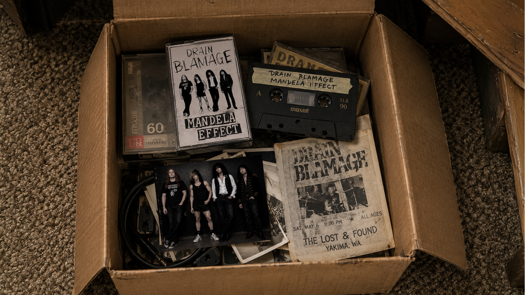 An open cardboard box containing a Drain Blamage cassette, band photograph, show flyer, and old music belongings